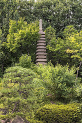 Stone tower pagoda in front of the central pond of Mejiro Garden which is surrounded by large flat stones under the foliage of  the Japanese pines trees and a variety of momiji maple trees.