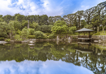 Obraz premium Hexagonal Gazebo in the central pond of Mejiro Garden where ducks are resting and which is surrounded by large rocks and stone pagoda under the foliage of the pines trees and Momiji maple trees.