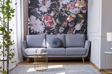 Sunlit, gray sofa by a floral print wall in the nook of a feminine living room interior with golden...