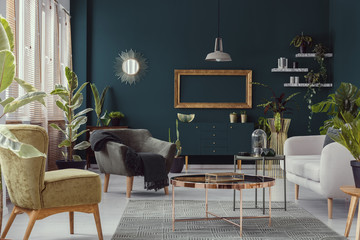 Copper table between armchairs and couch in green flat interior with plants and mirror. Real photo
