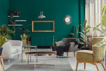 Armchairs and sofa next to copper table in green apartment interior with mockup of frame. Real photo