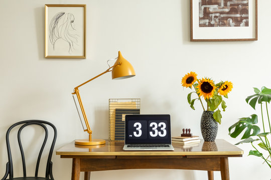 Sunflowers, yellow lamp and laptop on wooden desk in home office interior with posters. Real photo