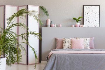 Pink pillows on grey bed in pastel bedroom interior with palm and poster on bedhead. Real photo