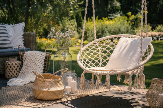 A beige string swing with a pillow on a patio. Wicker baskets, a rug and a blanket on a wooden deck in the garden.