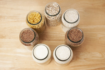 Various uncooked cereals, grains, and pasta for healthy cooking in glass jars on wooden table, white background, horizontal composition. Clean eating, balanced dieting food