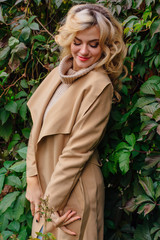 Young beautiful woman in sweater and coat stands next to the background of wild grapes leaves