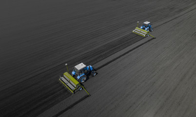 Aerial view of two blue tractors plows the earth in field on a summer day against a black earth background. Agriculture. Two tractors travel one after another along the black field