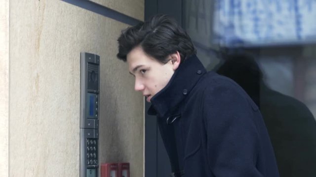 Young teenager talking on intercom by apartment building entrance
