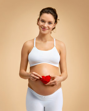 Happy pregnant woman holding red heart near her belly on beige background. Pregnancy, maternity, preparation and expectation concept