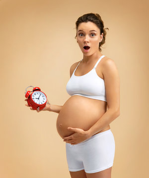Shocked pregnant woman holding alarm clock on beige background. Pregnancy, maternity, preparation and expectation concept