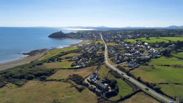 Aerial view, pan move. Drone panorama of sea with silhouette of Criccieth castle, grass fields, road and town in background