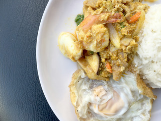 Stir fry prawns and squid with curry power thai style with fried egg with rice in white dish on black fabric texture background. Thai style food.