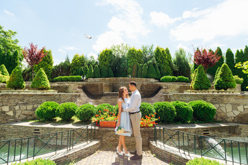 The guy embraces his girlfriend and they look at each other against the background of a park with beautiful landscape design.