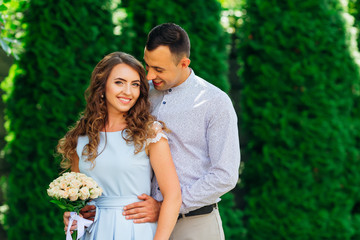 beautiful girl with an elegant hair style holds a bouquet of flowers and looks at the camera lens and her boyfriend hugs behind her waist
