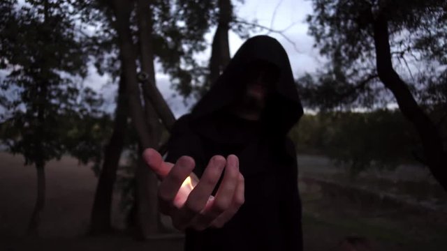 a fire mage in a black raincoat outdoors with a sword behind his back. taming the fire with your hand.