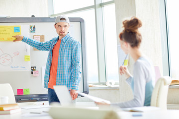 Obraz premium Confident handsome young guy with beard standing at whiteboard with schemes and sticky notes and presenting monthly plans at meeting