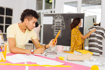 Drawing sketches. Attentive creative tailor thoughtfully looking at the screen of his device while drawing sketches for the next type of clothes