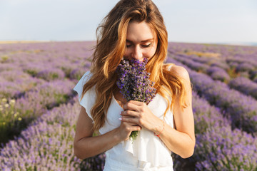 Photo of caucasian young woman in dress holding bouquet of flowers, while walking outdoor through lavender field in summer - obrazy, fototapety, plakaty