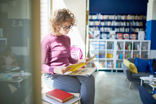 Female student with curly hair and eyeglasses preparing her report, she is sitting on window siil with book at library