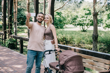 mother and father taking selfie with smartphone near baby carriage in park