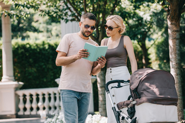 parents standing near baby carriage in park and reading book