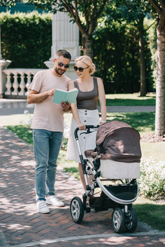 parents standing with baby carriage in park and looking at book