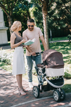 smiling parents standing near baby carriage in park and holding book