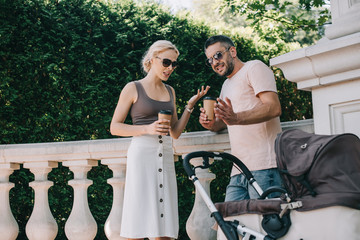 parents holding disposable coffee cups and quarreling near baby carriage in park