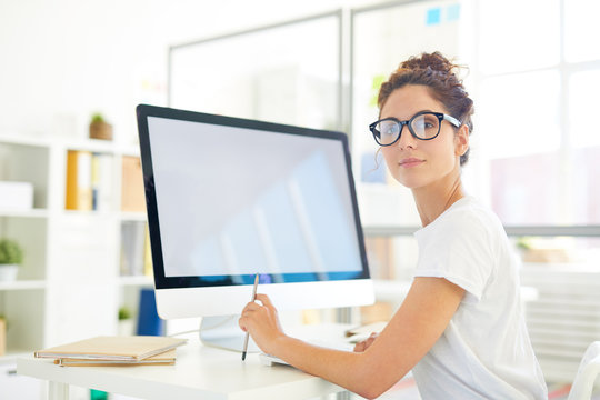 Young professional designer looking at you while sitting by desk in front of computer screen