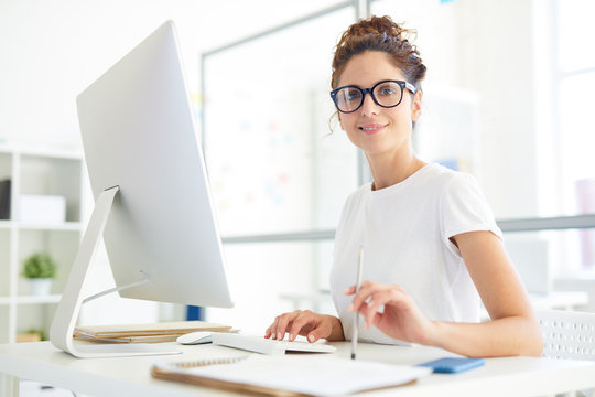 Young confident manager or designer sitting by her workplace in office in front of computer monitor