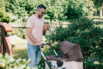 father walking with baby carriage in park and holding coffee in paper cup