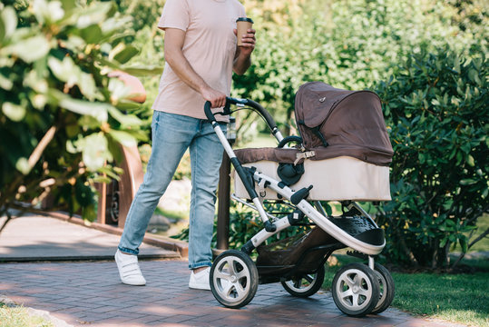 cropped image of father walking with baby carriage and disposable coffee cup in park