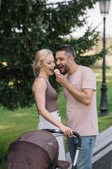 smiling husband feeding wife with ice cream near baby carriage in park