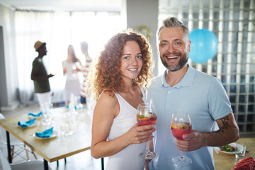 Young cheerful couple with drinks in wineglasses cheering up while looking at camera at party