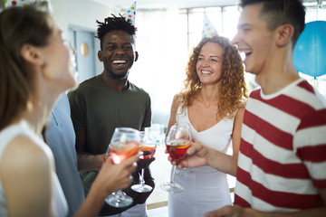 Happy laughing intercultural friends with drinks having fun while cheering up at home party
