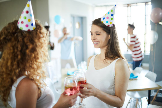 Cheerful birthday girls toasting with drinks in wineglasses at home party with dancing guys on background