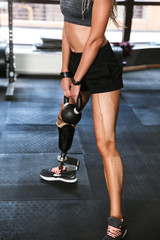 Naklejka na ściany i meble Portrait of fitness disabled woman with prosthesis in sportswear doing crossfit exercises, and lifting dumbbell in gym