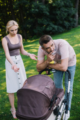 father smoking cigarette near baby carriage in park, angry mother gesturing and looking at him