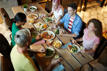 Group of five young people in casualwear sitting by wooden table, talking and enjoying healthy...