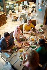 Five young friends sitting by large served wooden table and enjoying food and talk in cafe or restaurant