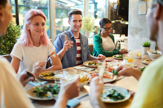 Happy guy pointing at one of his friends by dinner in cafe while talking to her