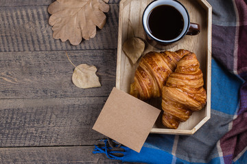 Autumn composition. Black coffee with croissants and empty craft paper card on tray, plaid and autumn leaves on a wooden background. Flat lay, top view, copy space.