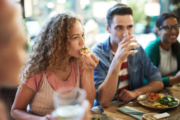 Hungry casual girl eating pizza by dinner among her friends and listening to one of them during chat