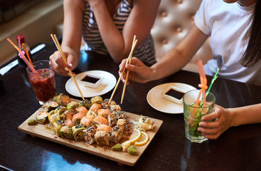 Fototapeta na wymiar Two yang girls are eating delicious rolling sushi at restaurant served on the wooden board with chopsticks, soy sauce and cocktails