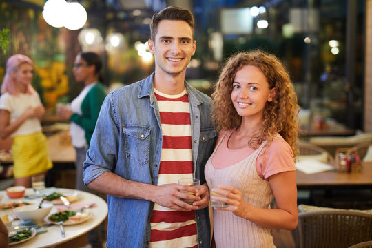 Happy young man and woman with drinks looking at you while enjoying party with friends in cafe
