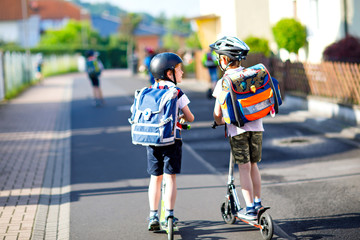 Two school kid boys in safety helmet riding with scooter in the city with backpack on sunny day....