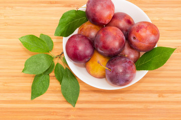 Plums in white bowl and leaves on a wooden surface