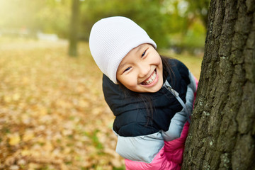 Laughing adorable girl in white beanie and striped jacket looking out of tree trunk in park
