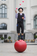 The clown and balancer juggles with pink balls , standing on a big red ball in the street of a European city - 219771390
