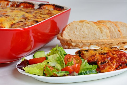 Baked layers of aubergine, cheese and tomato topped with melted cheese served with salad and bread with the remainder of the baked aubergine in a red ceramic baking dish to the rear.